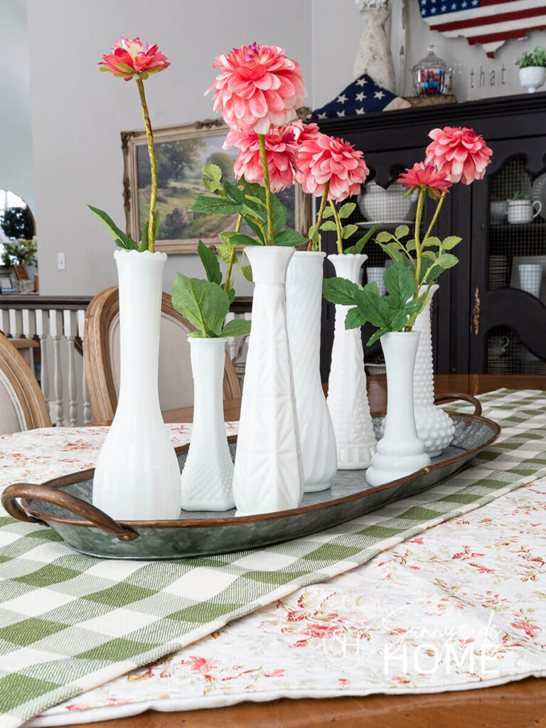 Decorating the dining table for summer with a floral tablecloth and a green gingham table runner, topped with a centerpiece of milk glass bud vases in a galvanized and copper tray and filled with pink zinnias.