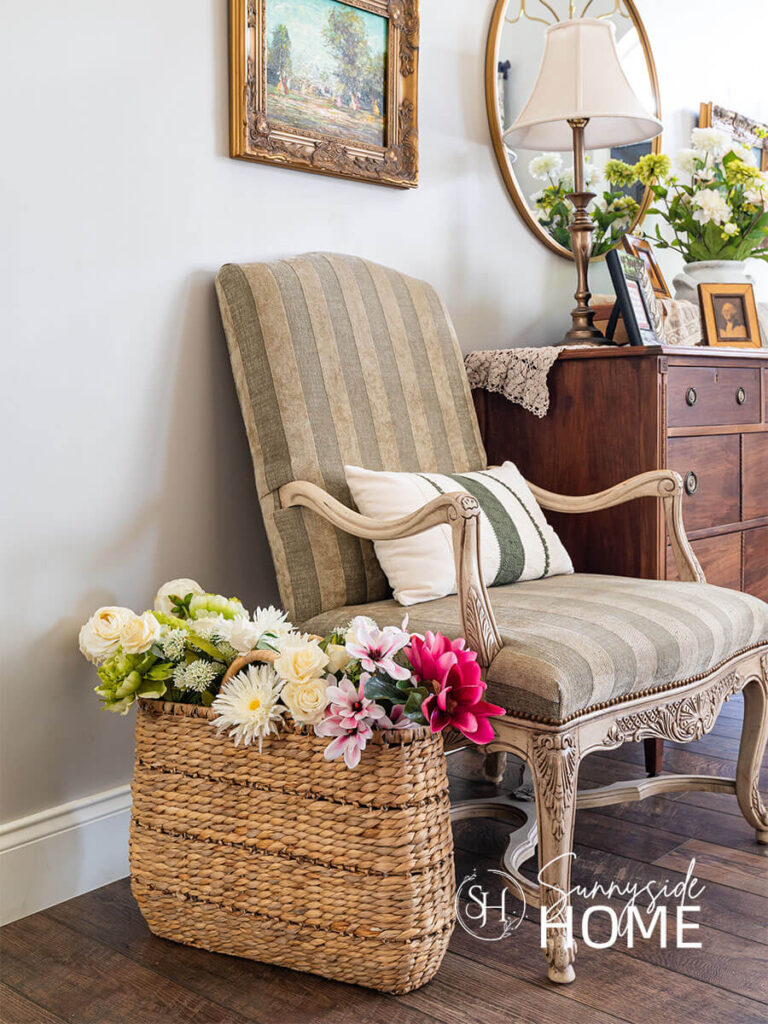 Decorating ideas for summer, large basket filled with a variety of flowers in shades of white, light greens and pinks, placed on the floor next to a green striped vintage chair with lumbar pillow in the entryway.