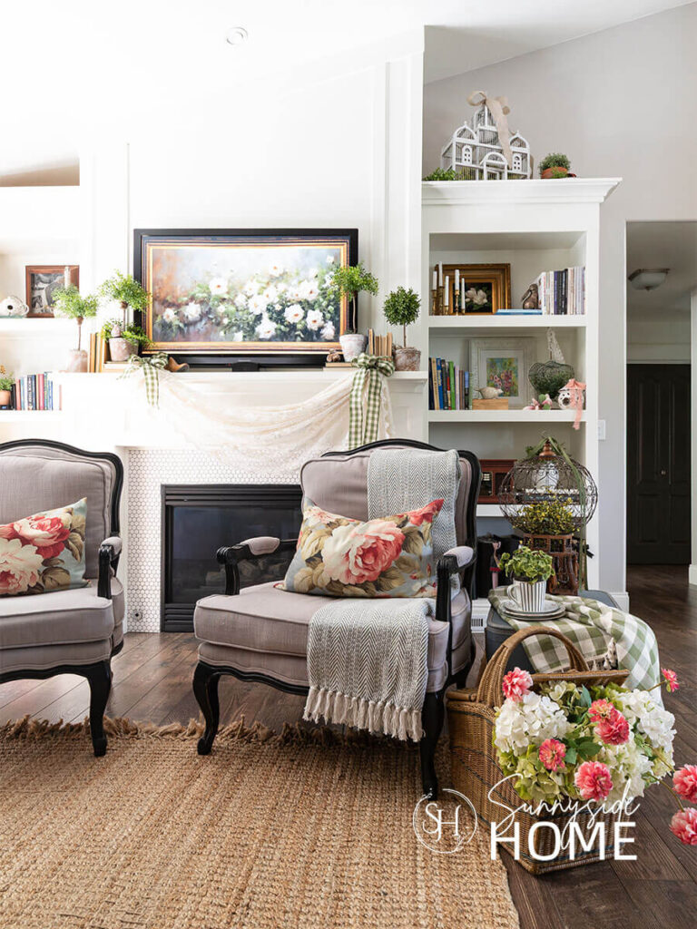 Decorating ideas for summer in the family room. A pair of chairs styled with floral lumbar pillows and a cotton throw in front of the mantel. The mantel is decorated for summer with a vintage lace table swag and embellished with green gingham bows. A. top the mantel are topiary and stacked vintage books.