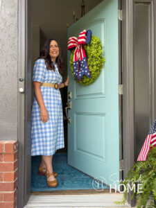 Woman open front door, on the door is an boxwood wreath embellished with a red, white and blue patriotic bow.