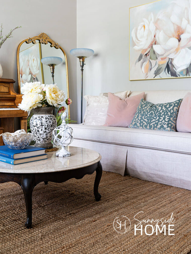 Decorating ideas for the living room. Large floral art is hung above the sofa. The round marble coffee table is styled with a large black and white vase filled with giant cream peonies, bird decor, book stack topped with a vintage crystal bowl filled with vintage doilies. The linen sofa is embellised with blue, blush and off whtie pillows.