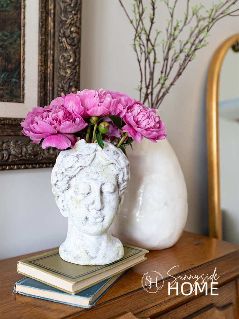 A aged woman's head vase is filled with pink peonies and placed on a vintage book stack and styled with a tall vase with branches on the piano.