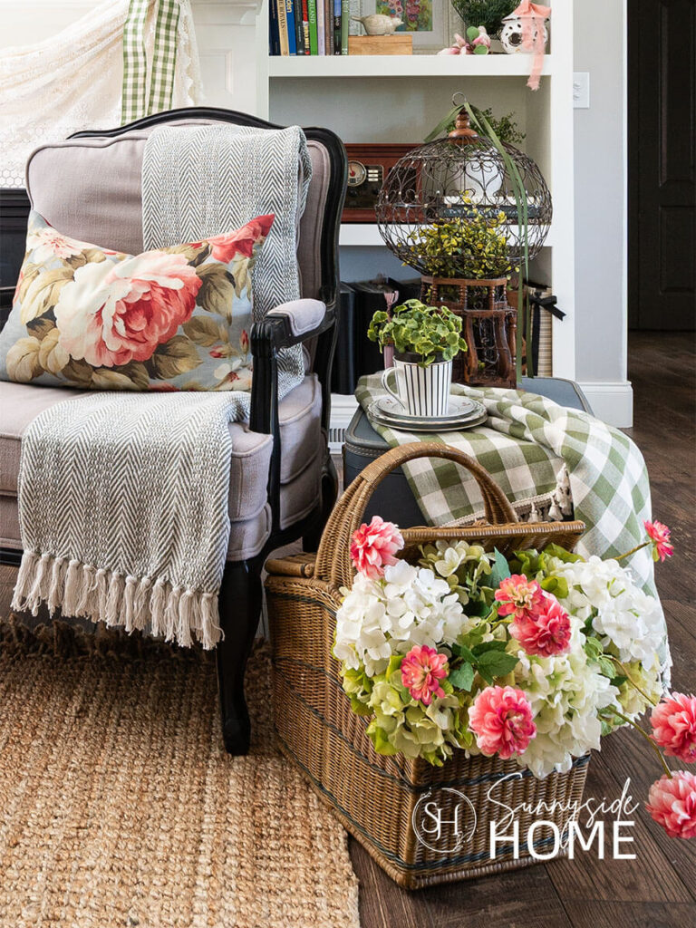 Decorating ideas for summer in the living room. A stack of vintage suitcases styled with a green gingham tablecloth, plate stake and teacup with a plant and a victorian birdcage with greenery and ribbon used as a side table next to a chair. A vintage picnic basket is filled with faux hydrangeas and pink zinnias.