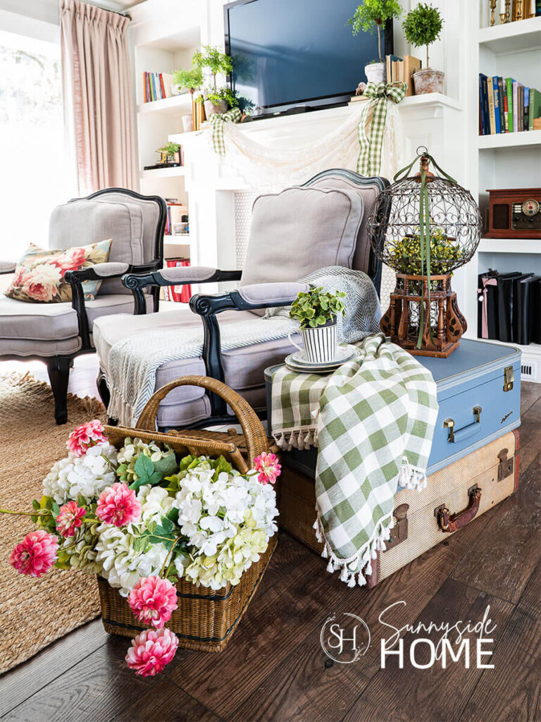 Decorating ideas for summer in the living room. A stack of vintage suitcases styled with a green gingham tablecloth, plate stake and teacup with a plant and a victorian birdcage with greenery and ribbon used as a side table next to a chair. A vintage picnic basket is filled with faux hydrangeas and pink zinnias.