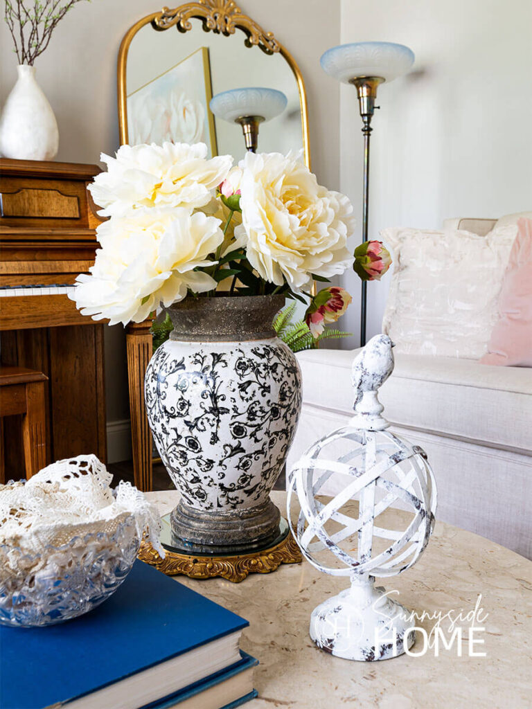 Decorating ideas for summer in the living room. The round marble coffee table is styled with a large black and white vase filled with giant cream peonies, bird decor, book stack topped with a vintage crystal bowl filled with vintage doilies.