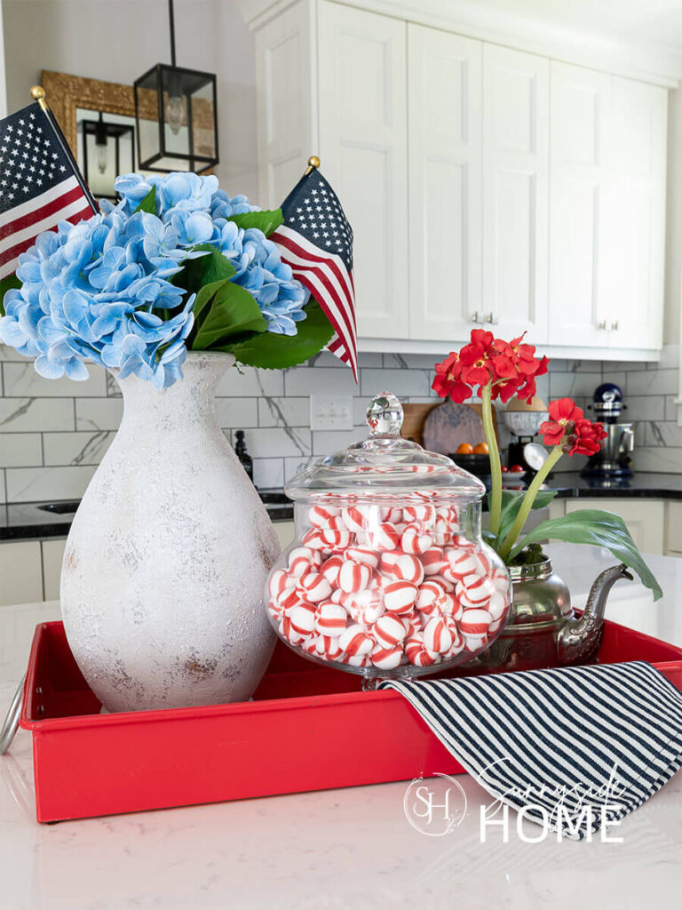 Decorating ideas for summer on the kitchen island. A red metal tray styled with a rustic pottery vase filled with blue hydrangeas and mini flags, apothecary gar will with peppermints and a vintage silver teapot with a red potted plant.