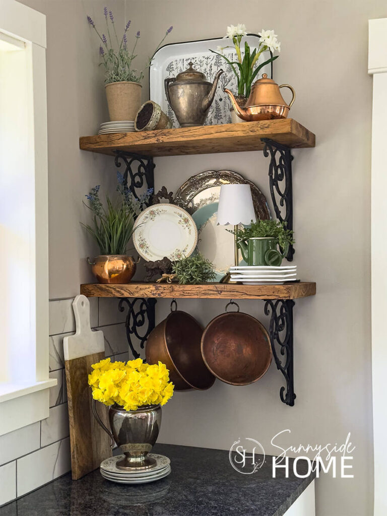 Open kitchen shelves are decorated for summer with several potted flowering plants, vintage floral plate on a metal stand, stacked plates as risers for teapots and plants.