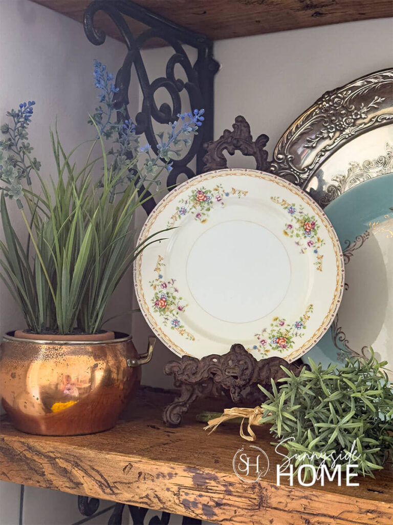 Kitchen shelf is decorated for summer with a plant in a vintage copper pot, vintage floral plate, silver tray and blue dinner plate.