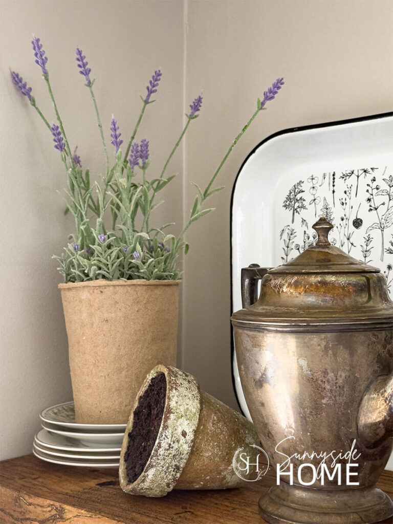 A potted lavender plant on stacked vintage plates, rustic clay pot, tarnished teapot and a black and white floral tray.
