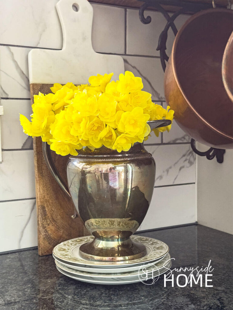 A silver pitcher with yellow flowers on a stack of thrifted plates on the kitchen.