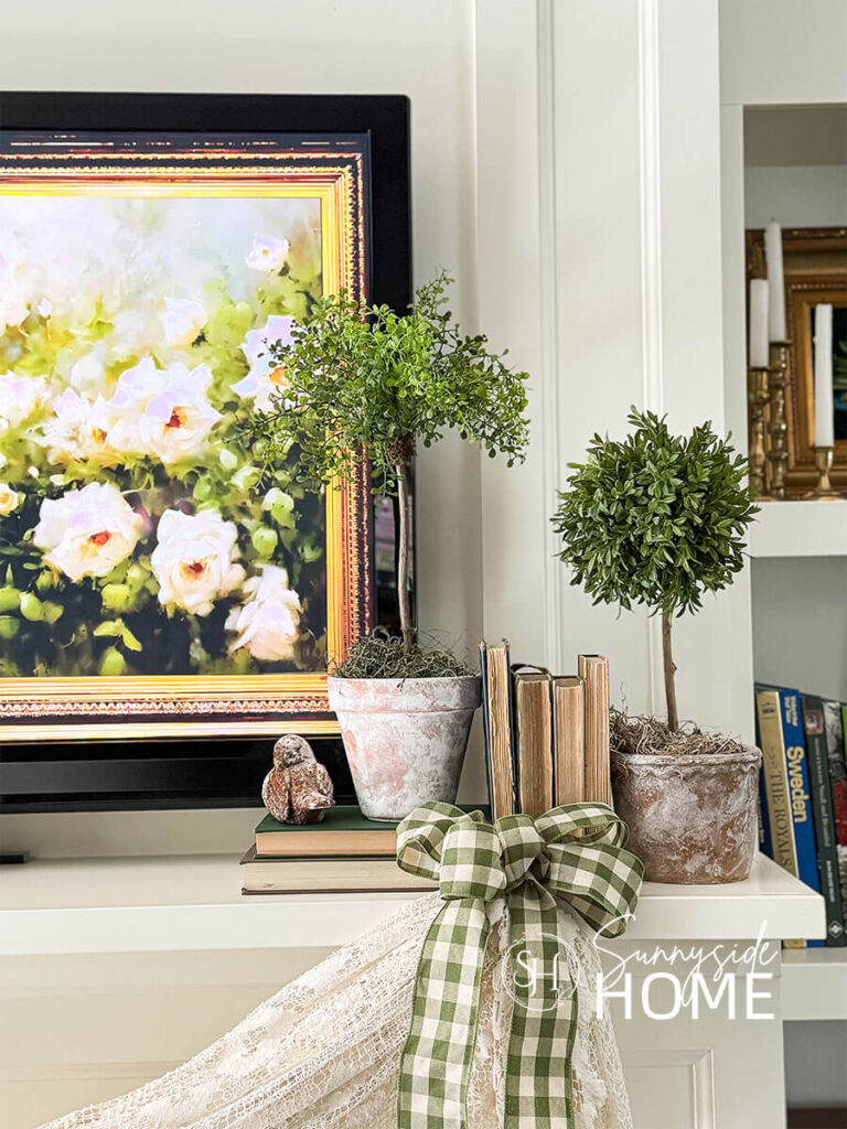 Decorating ideas for summer on the mantel. Topiaries styled with stacked vintage books and birds, a vintage lace tablecloth is swaged across the mantel and embellished with green gingham bows.
