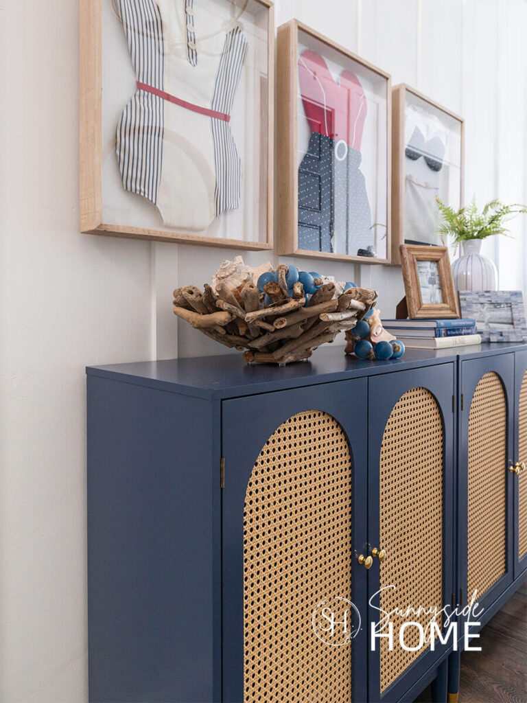 Entryway is decorated for summer framed red, white and blue vintage style swimsuits. The blue cabinet is styled with a driftwood bowl filled with seashells and a blue glass bead garland.