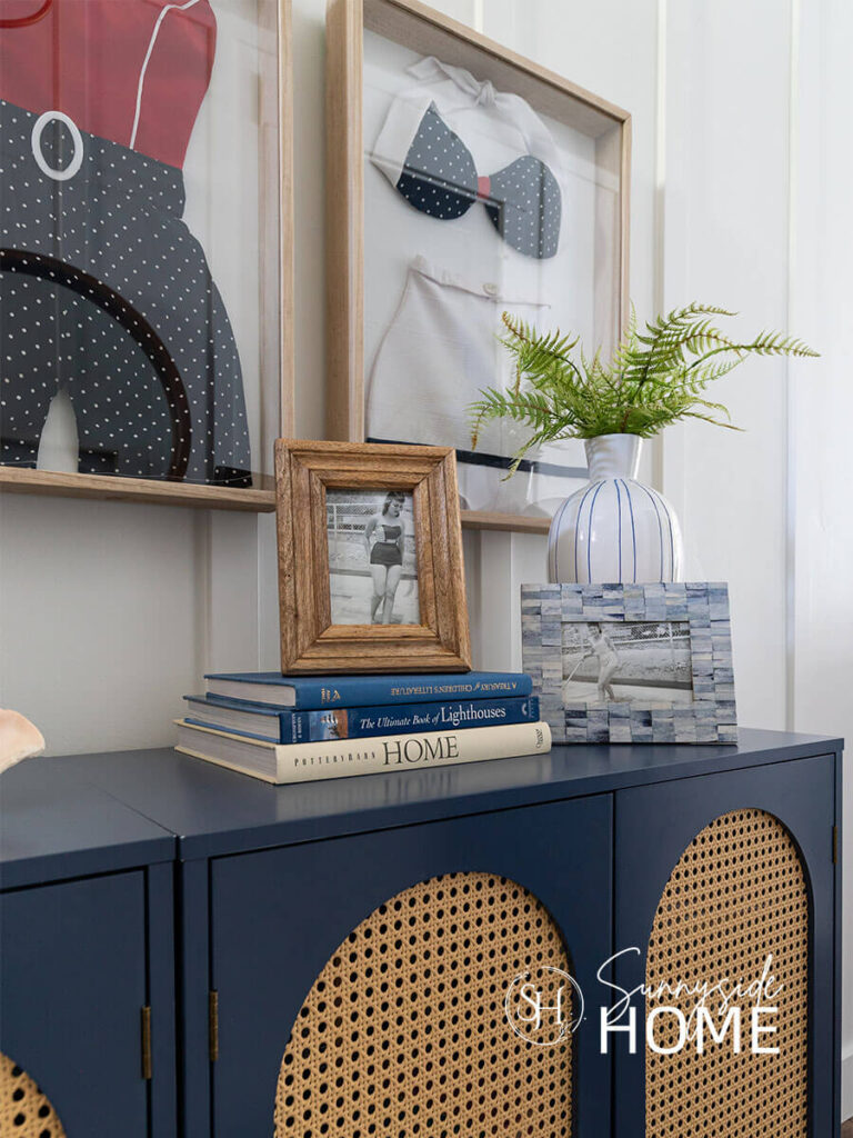 Blue arched cabinet is styled with a stack of blue and white books, blue and white vase with greenery and two vintage framed black and white photos of woman in swimsuits.