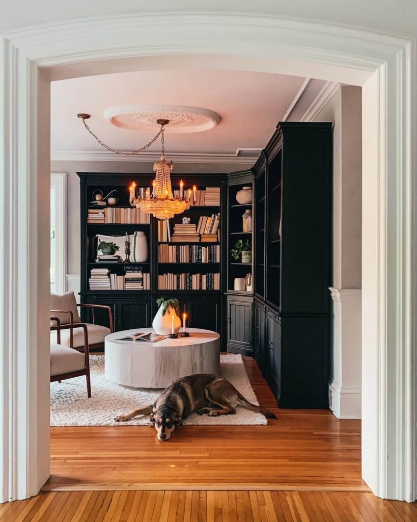 Black painted book shelves in an under used dining room with a modern round coffee table, chandelier and two reading chairs.