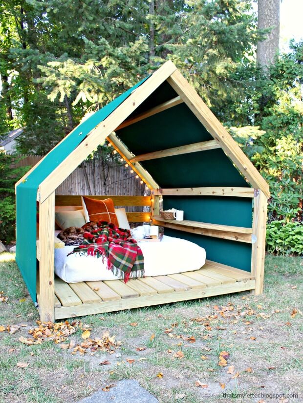 A DIY outdoor room is created with lumber and is draped with outdoor fabric and lights creating the perfect outdoor reading nook. Inside the framed room is a mattress covered with cozy linens and pillows.