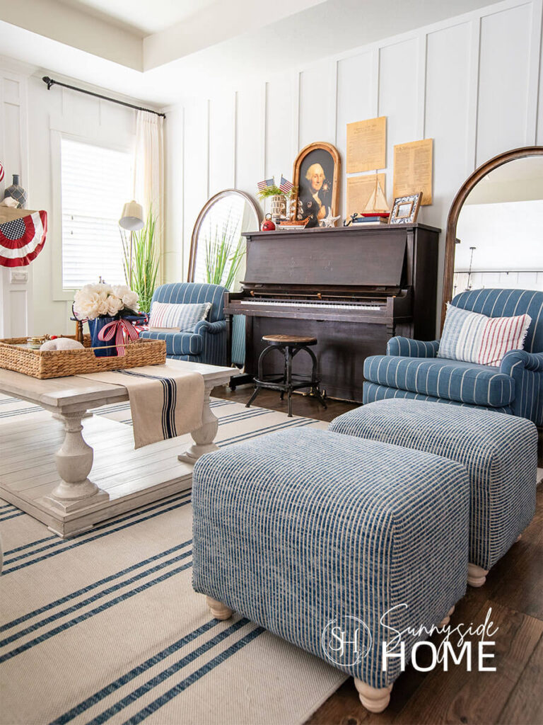 Summer decorating ideas with patriotic style in the living room: red, white and blue pillows on blue and white striped chairs on each side of the piano. Above the piano is a framed whimsical image of George Washington and patriotic documents. On the piano are sailboats, buoy, flags and a framed photos. Coffee table styling for summer in a large basket.