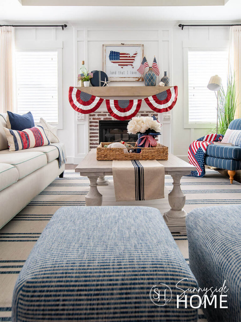 Fourth of July decorating ideas in the living room: coffee styled with blue and white table runner and a large basket tray filled with red, white and blue decor. The mantle is styled with a patriotic bunting, patriotic art above the mantle, and on the mantel are blue jugs, American flags, blue travel bag, jug of sand and a starfish.