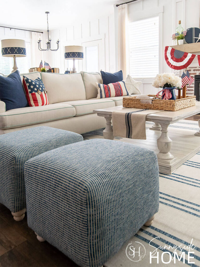 The sofa in the living room is decorated for the fourth of july with a variety of red, white and blue pillows. The white washed coffee table is styled with a blue and white table runner, topped with a hyacinth basket tray and red, white and blue decor.