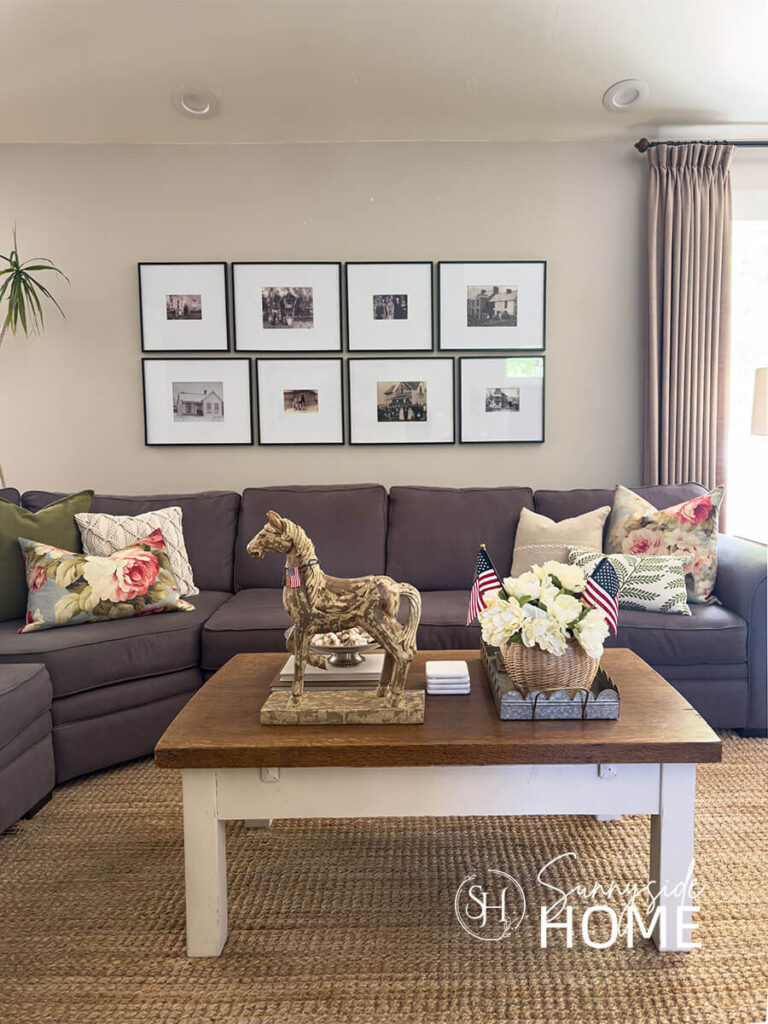 Sofa is decorated for summer with floral, linen and embroidered fern pillows. The coffee table is decorated for summer with a scalloped galvanized tray filled with a peony and flag bouquet in a wicker basket, horse statue, and footed bowl with collected seashells.