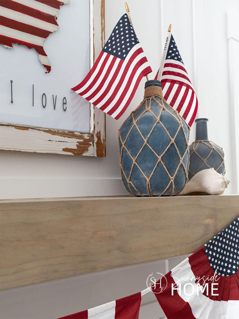 Two blue and jute jug vases are placed on the mantel with a large seashell and a pair of American flags.