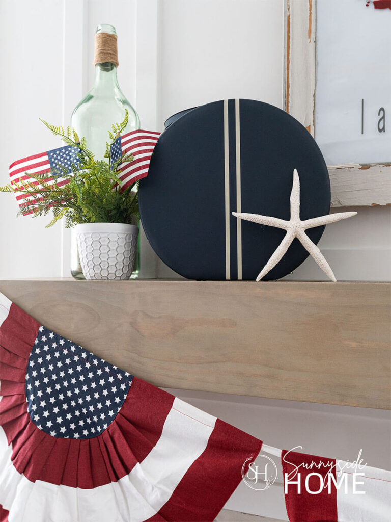 A round blue and white striped travel case is styled on the mantle with a starfish, potted fern with a pair of American flags and a green glass bottle.