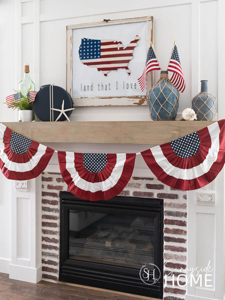 The mantle is styled with a patriotic bunting, patriotic art above the mantle, and on the mantel are blue jugs, American flags, blue travel bag, jug of sand and a starfish.