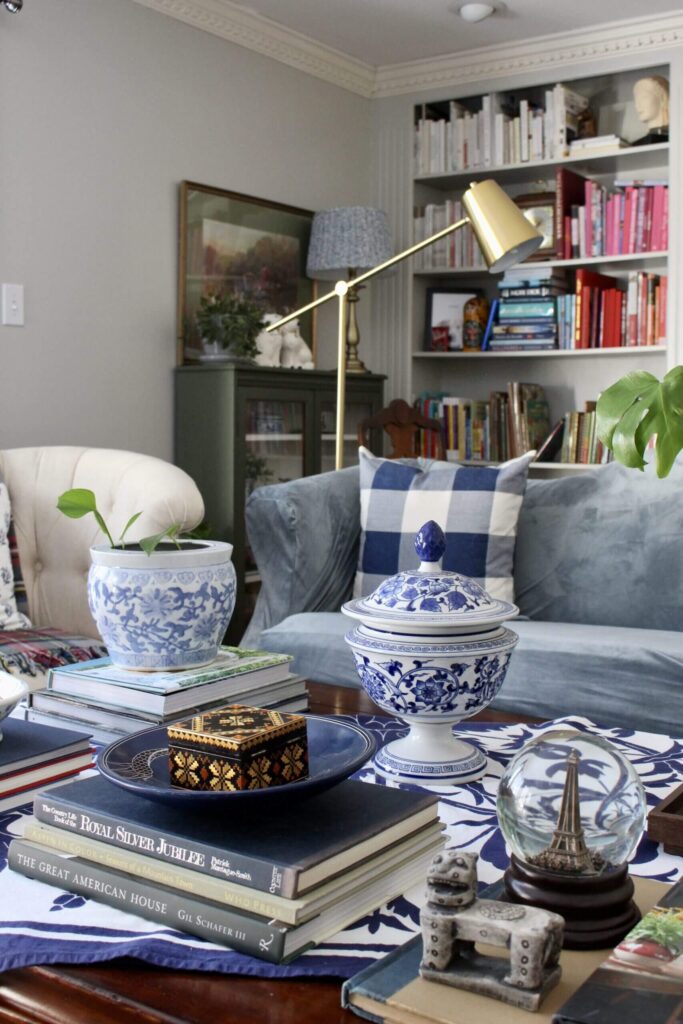 A cozy reading room with a sofa, chair, floor lamp, bookshelves and stacks of books on a coffee table.