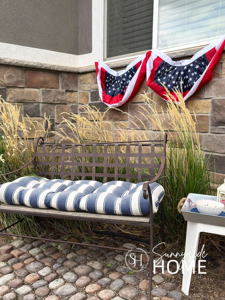 Fourth of July decorating ideas for outdoors, below the front window is a pair of patriotic buntings, a blue and white stripped cushion on a rustic bench.