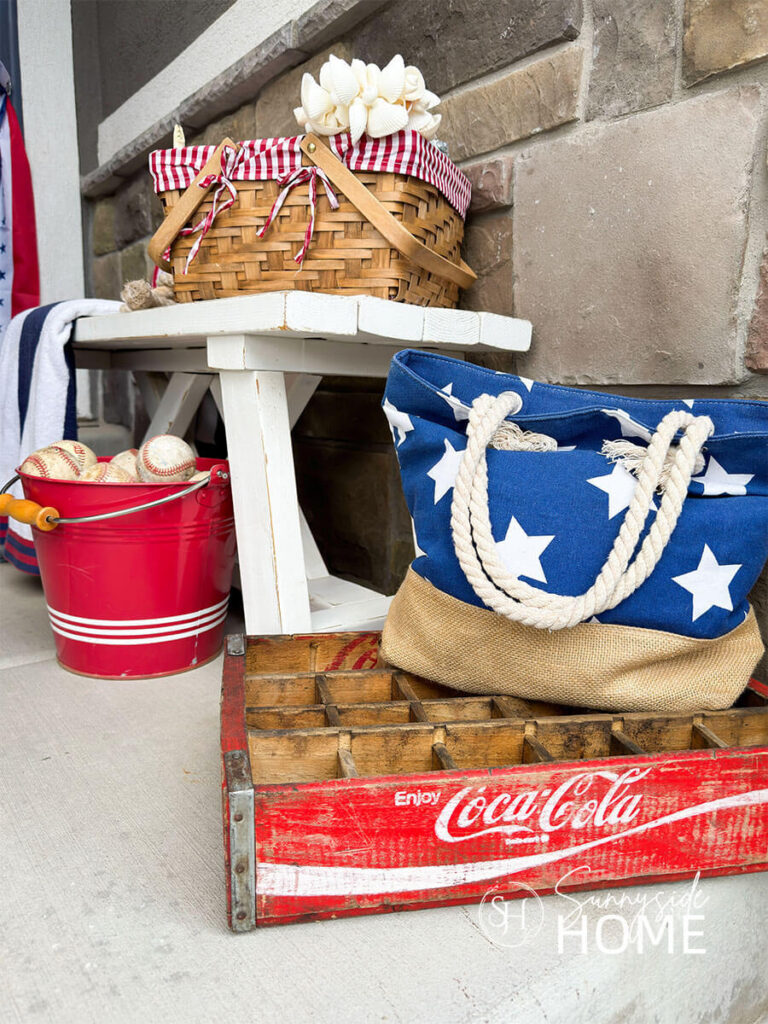 Fourth of July decorating ideas for the front porch, a white farmhouse bench is styled with a basket with a red and white gingham liner filled with seashells, under the bench if a red bucket filled with baseballs, a red coke crate topped with a blue beach back with white stars.