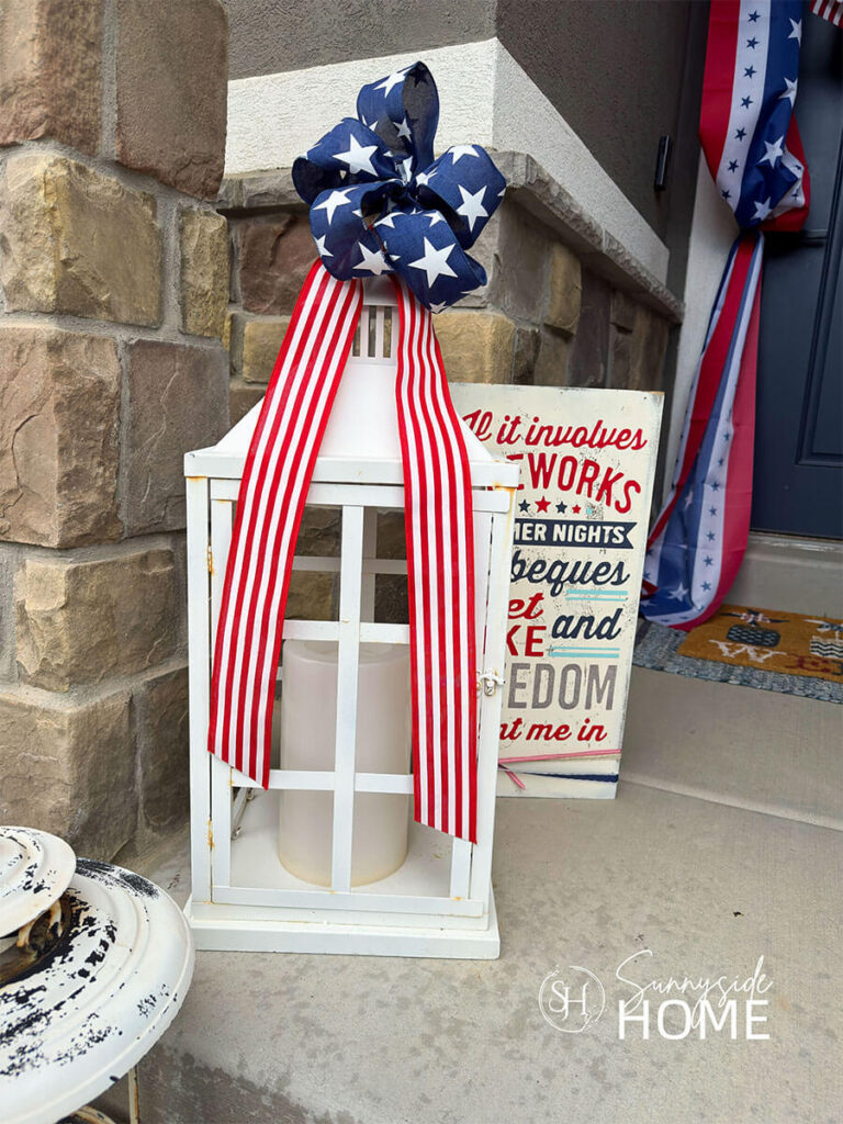 A white lantern is embellished for the 4th of July with a red and white striped ribbon and toped with a 5 loop bow made with navy blue ribbon with white stars.