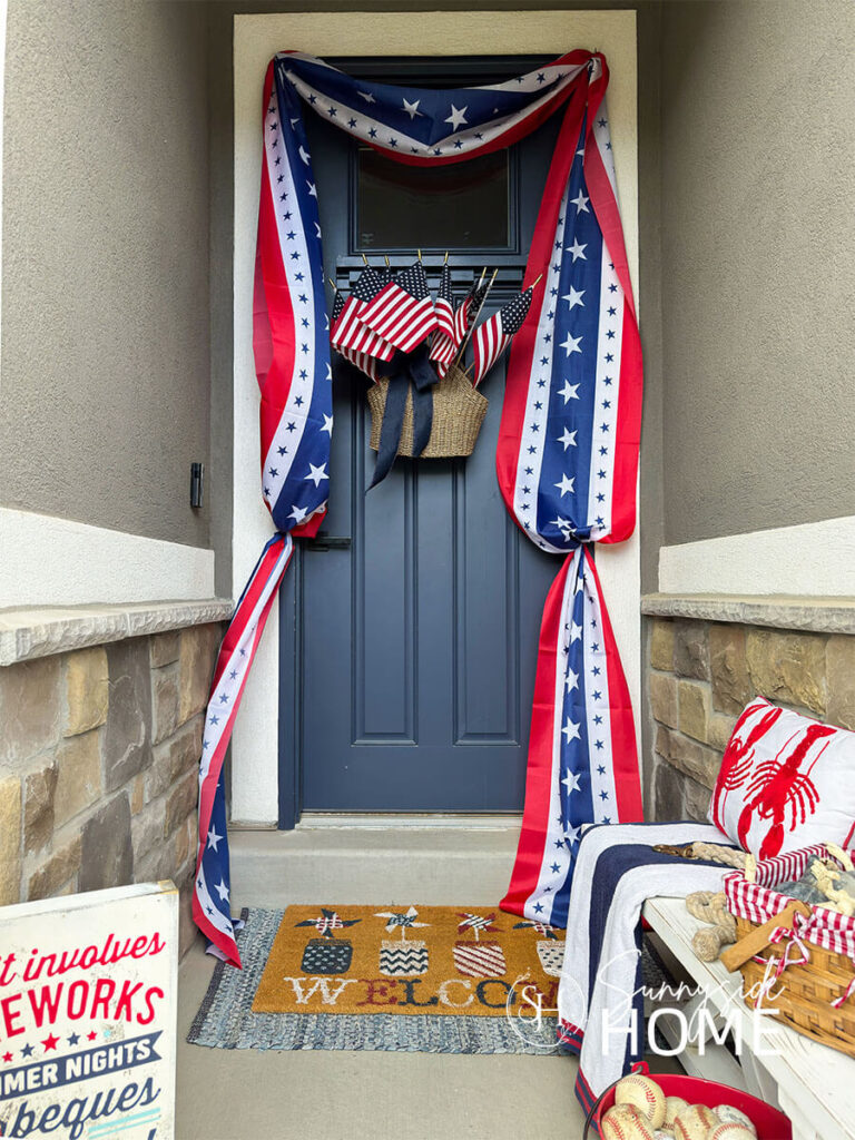 A festive Fourth of July decorated front porch, a navy blue door with a basket filled with American flags and a patriotic bunting hung around the door frame. A patriotic welcome mat layered over a denim rag rug. A white farmhouse bench is styled with a red, white and blue beach towel topped with a red lobster pillow.