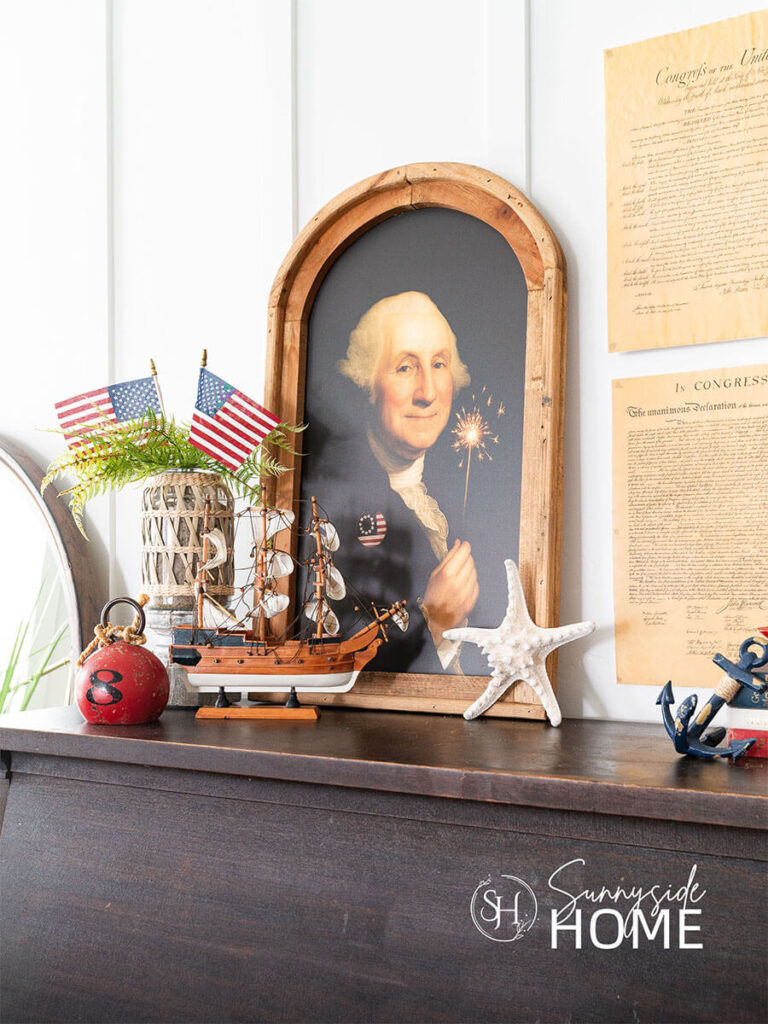A framed whimsical picture of George Washington is styled above the piano for the fourth of July with a sailboat, red buoy, starfish and a coastal vase with a fern and two American flags.