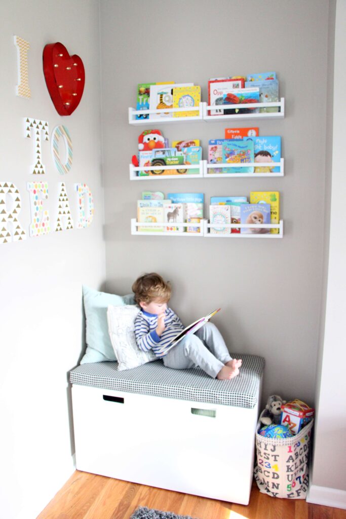 A corner of a kids playroom is dedicated to reading with wall mounted shelves with picture books and a toy chest topped with a cushion.