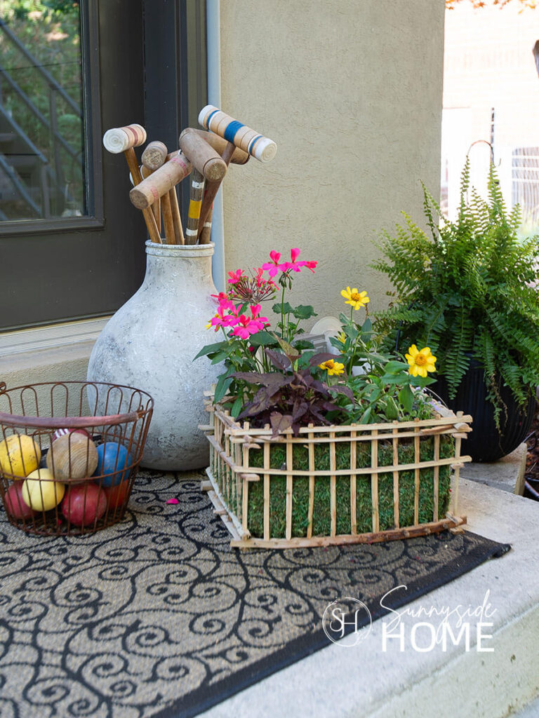 A backyard porch is styled with a thrifted wood basket filled with pink and yellow flowers along with a large jug filled with croquet mallets an egg basket filled with balls.