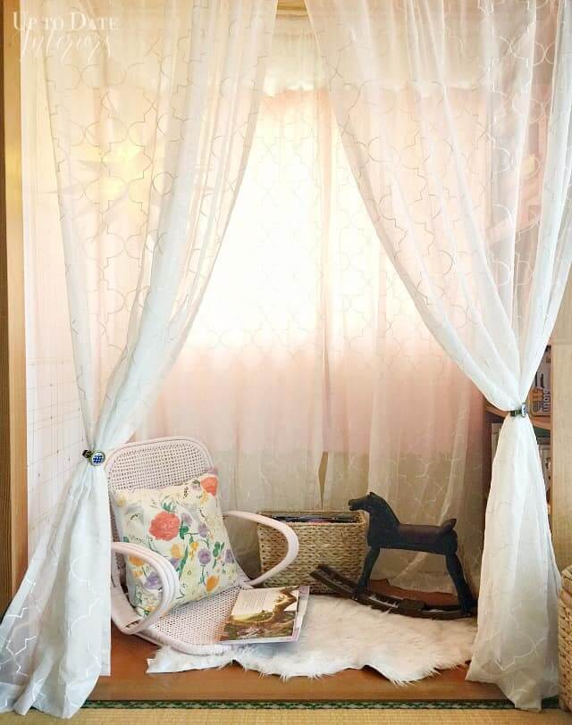 Lace curtains are hung to partition off a section of a room with creating a cozy reading nook with a floor chair, cozy rug and bookshelves.
