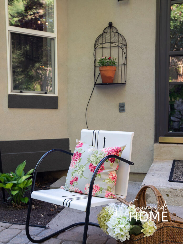 A wire birdcage shelf hung on a house styled with a clay pot with flowers. A painted vintage chair with a floral pillow and vintage picnic basket with hydrangea stems styled on a paver patio.