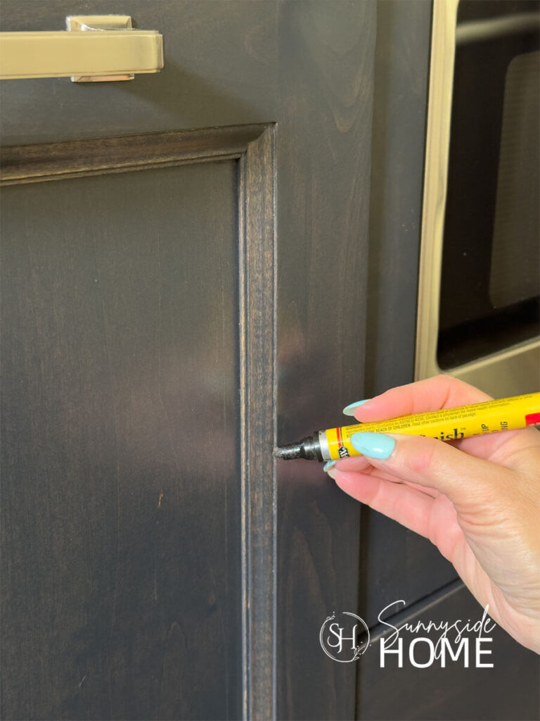 Woman applies stain to fix chip in finish on a dark stained kitchen cabinet door with a stain pen.