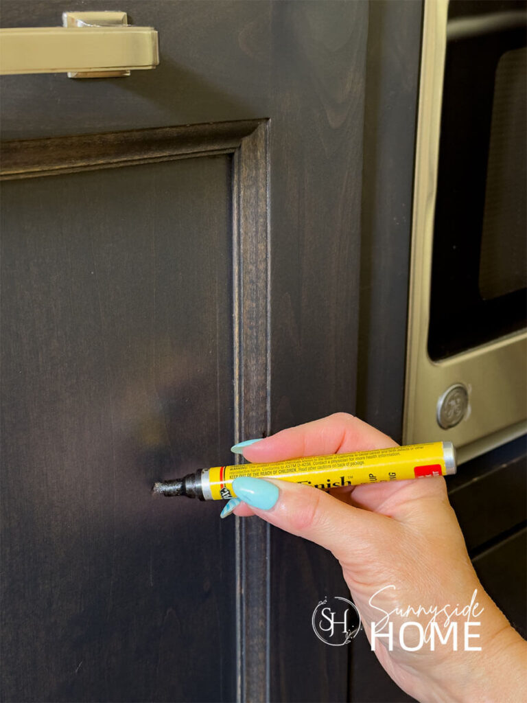 Woman applies stain to fix chip in finish on a dark stained kitchen cabinet door with a stain pen.
