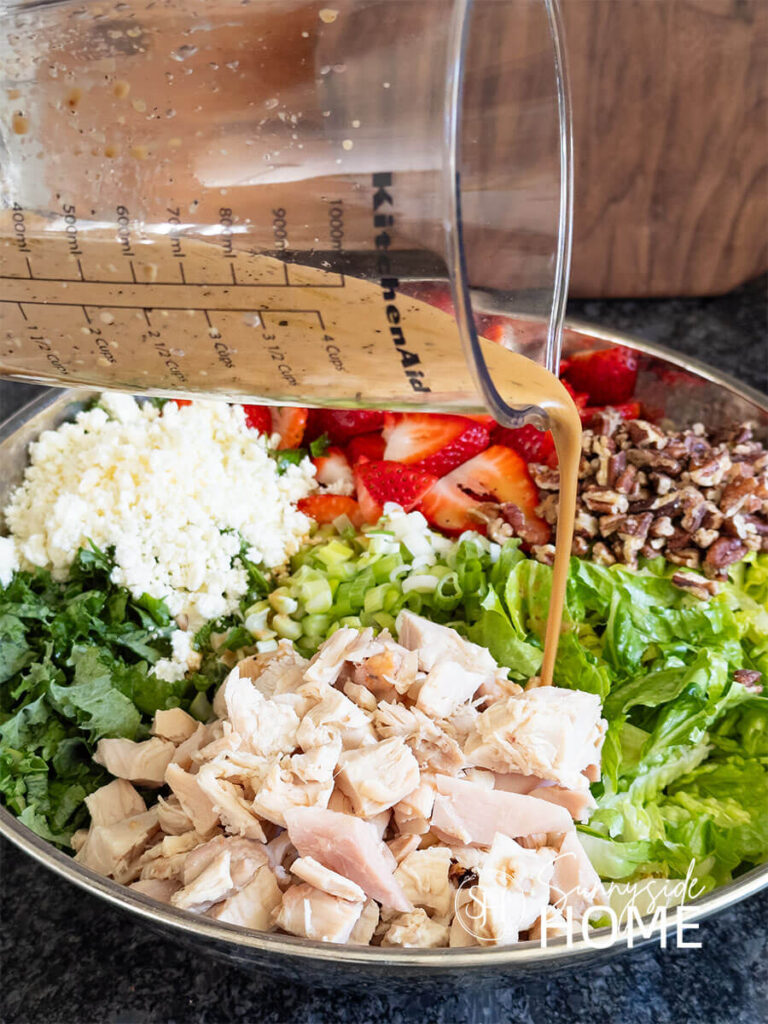 Lime maple balsamic vinaigrette is poured over the ingredients of mixed greens, chopped chicken, feta cheese, sliced strawberries, candied pecans and green onions in a silver serving bowl.
