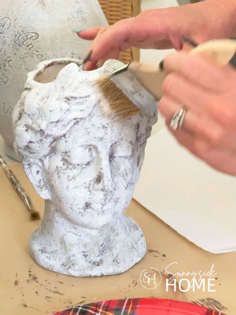 A woman dry brushes on white paint to achieve a worn and weathered finish to a head vase.