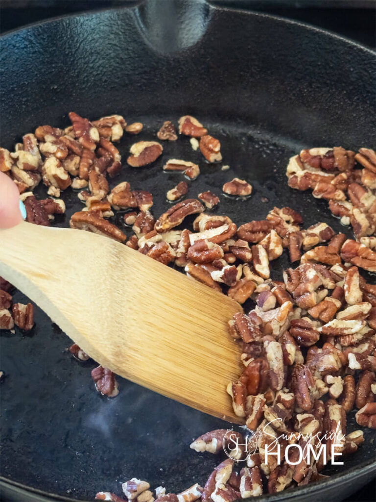Woman tosses the toasted pecans in a cast iron pan with a wood spatula.