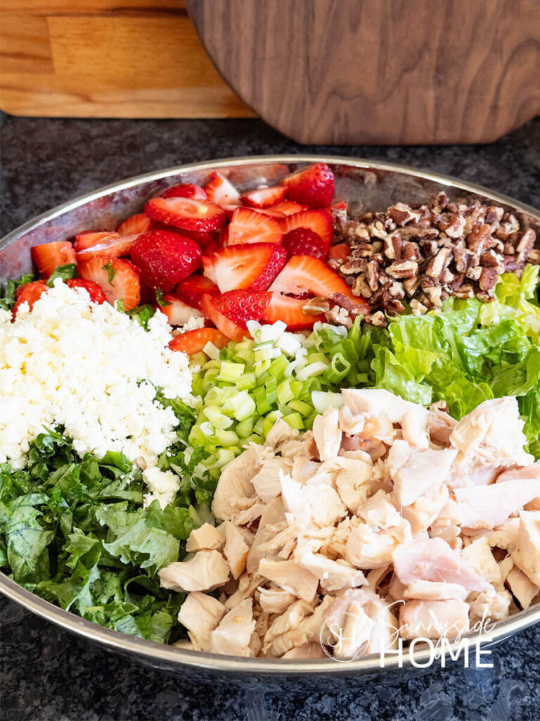 Salad ingredients in a silver bowl. The salad features a mix of greens, savory chicken, sweet fresh strawberries, green onion, crunchy pecans, creamy feta cheese. A healthy summer salad.