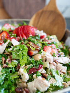 Strawberry chicken salad in a silver bowl with wooden serving spoons. The salad features a mix of greens, savory chicken, sweet fresh strawberries, green onion, crunchy pecans, creamy feta cheese and a sweet and tangy lime maple balsamic vinaigrette. A healthy summer salad.