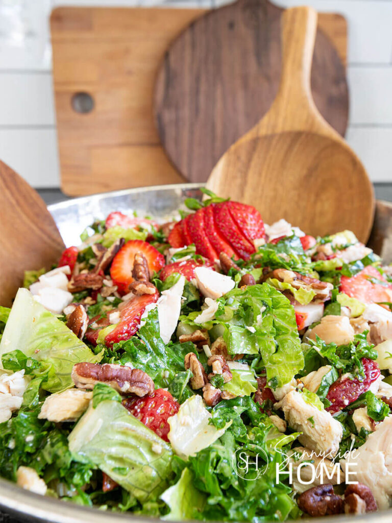 Strawberry chicken salad in a silver bowl with wooden serving spoons. The salad features a mix of greens, savory chicken, sweet fresh strawberries, green onion, crunchy pecans, creamy feta cheese and a sweet and tangy lime maple balsamic vinaigrette. A healthy summer salad.
