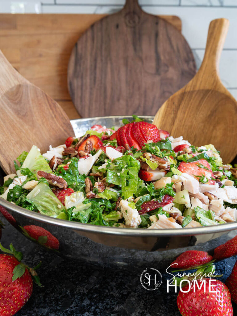 Strawberry chicken salad in a silver bowl with wooden serving spoons. The salad features a mix of greens, savory chicken, sweet fresh strawberries, green onion, crunchy pecans, creamy feta cheese and a sweet and tangy lime maple balsamic vinaigrette. A healthy summer salad.
