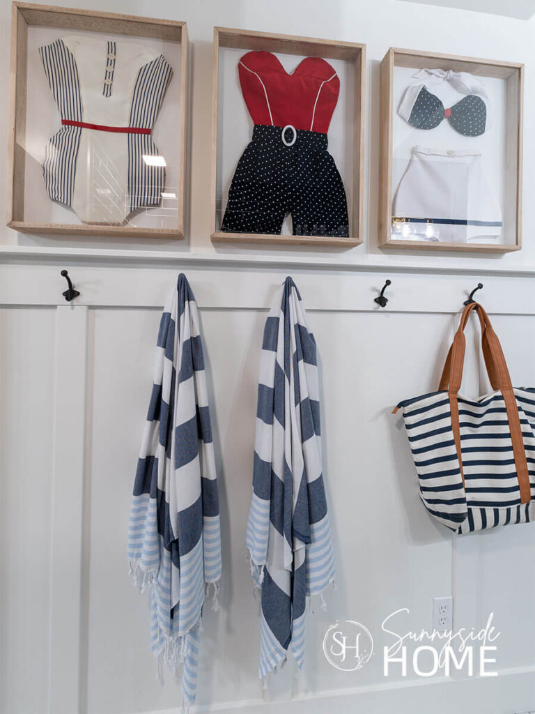 Easy decorate a bathroom with blue and white striped towels on wall hooks. Framed vintage inspired swimsuits are framed in shadow boxes above the row o hooks.