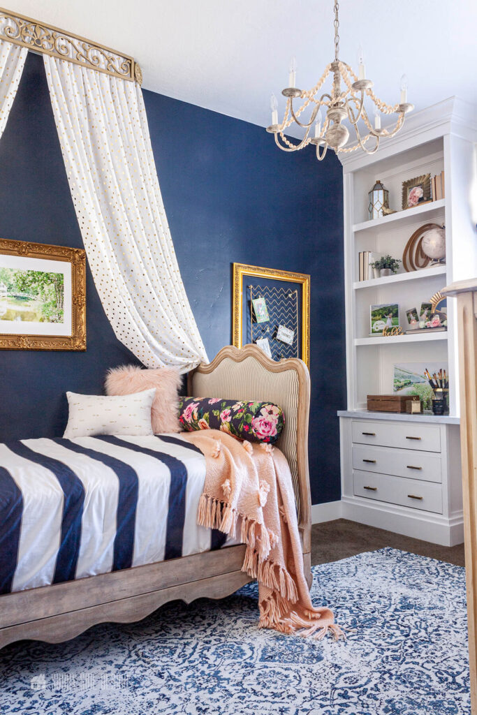 In a girls bedroom a navy blue wall anchors the room. A French day bed against the blue wall with a white canopy with a gold filigree crown. The bed is dressed with blue and white striped comforter and accented with pink pillows and throw.