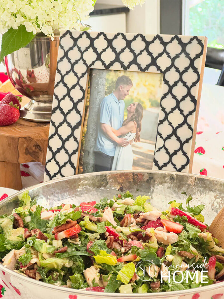 Bridal shower buffet with a strawberry table runner topped with a strawberry and chicken salad, photo of the bride and groom, floral arrangement with white hydrangeas and strawberries.