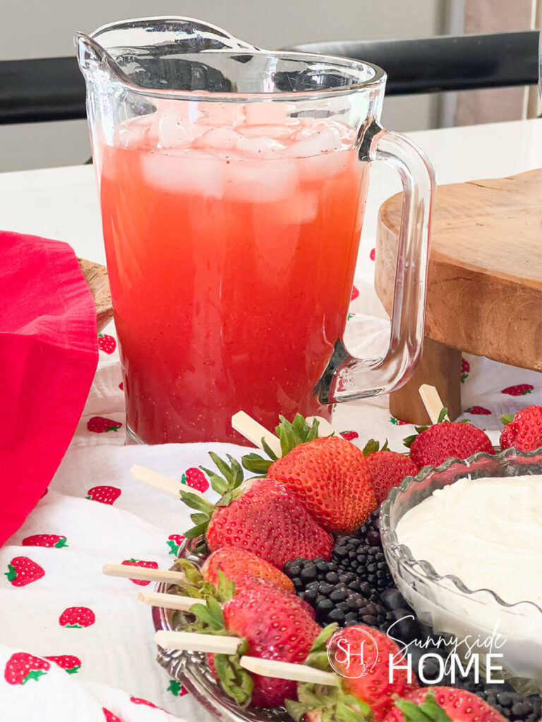 A pitcher of fresh strawberry lemonade placed on a strawberry table runner along with a tray of berry fruit kebabs and dip.