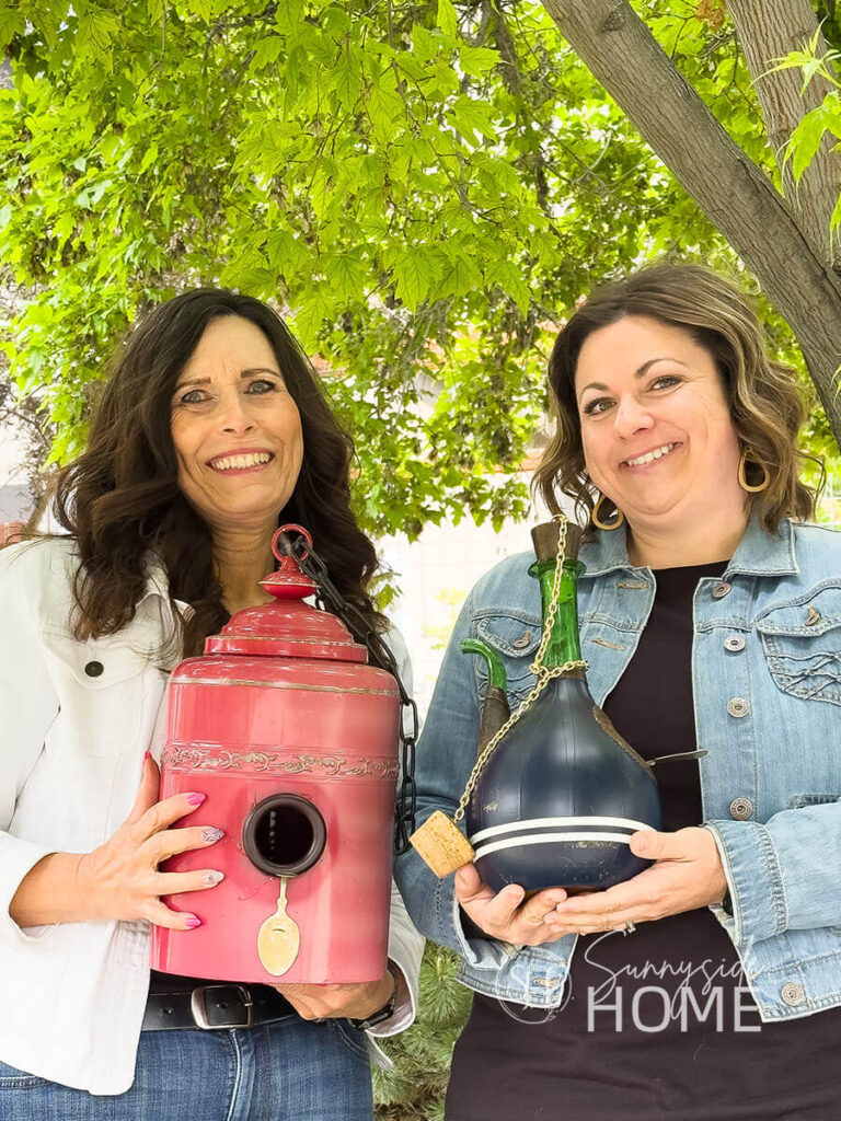 Two women hold their upcycled thrifted birdhouses. One is made from a red canister and the second one is made from a wine chiller, painted navy blue with white stripes.